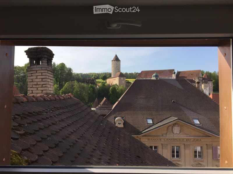 View from a window showing a historic town with red-tiled roofs, a prominent stone chimney, a tall tower, and a building labeled "BANQUE CANTONALE DU JURA" with classical architecture.