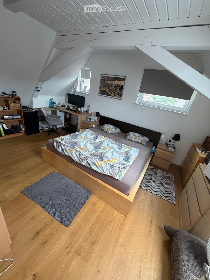 Cozy attic bedroom with wooden floors, slanted ceilings, and large windows. Features a bed with a leafy-patterned duvet, a desk with a computer, and built-in shelving.