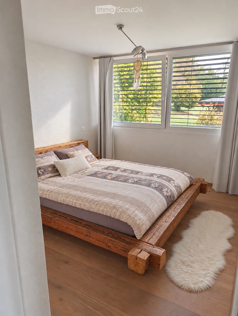 Cozy bedroom with a wooden bed, patterned bedding, and a fluffy white rug on a wooden floor. Large windows with blinds offer a view of greenery outside.