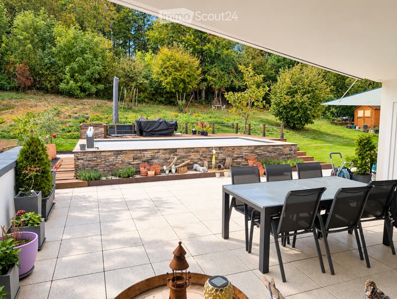 Outdoor patio with a dining table and chairs, surrounded by potted plants and a stone barbecue area, with a swimming pool and garden in the background.