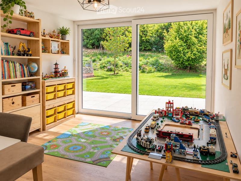 Brightly lit children's room with large windows overlooking a garden, featuring a play mat with a road design, a wooden table with a toy train set, shelves with books and toys, and a comfortable chair.