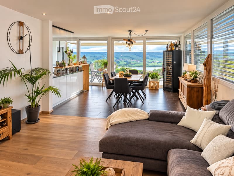 Open-plan living and dining area with wooden flooring, large windows offering scenic views, a grey sectional sofa, a coffee table, a dining table with chairs, a wine cooler, and several potted plants.