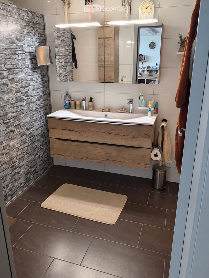 Modern bathroom with a wooden vanity, large mirror, stone accent wall, and various toiletries on the sink counter.