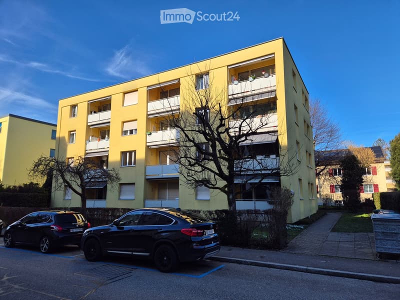 Multi-story residential building, yellow exterior, several balconies with railings, landscaped entrance, and parking area in front.