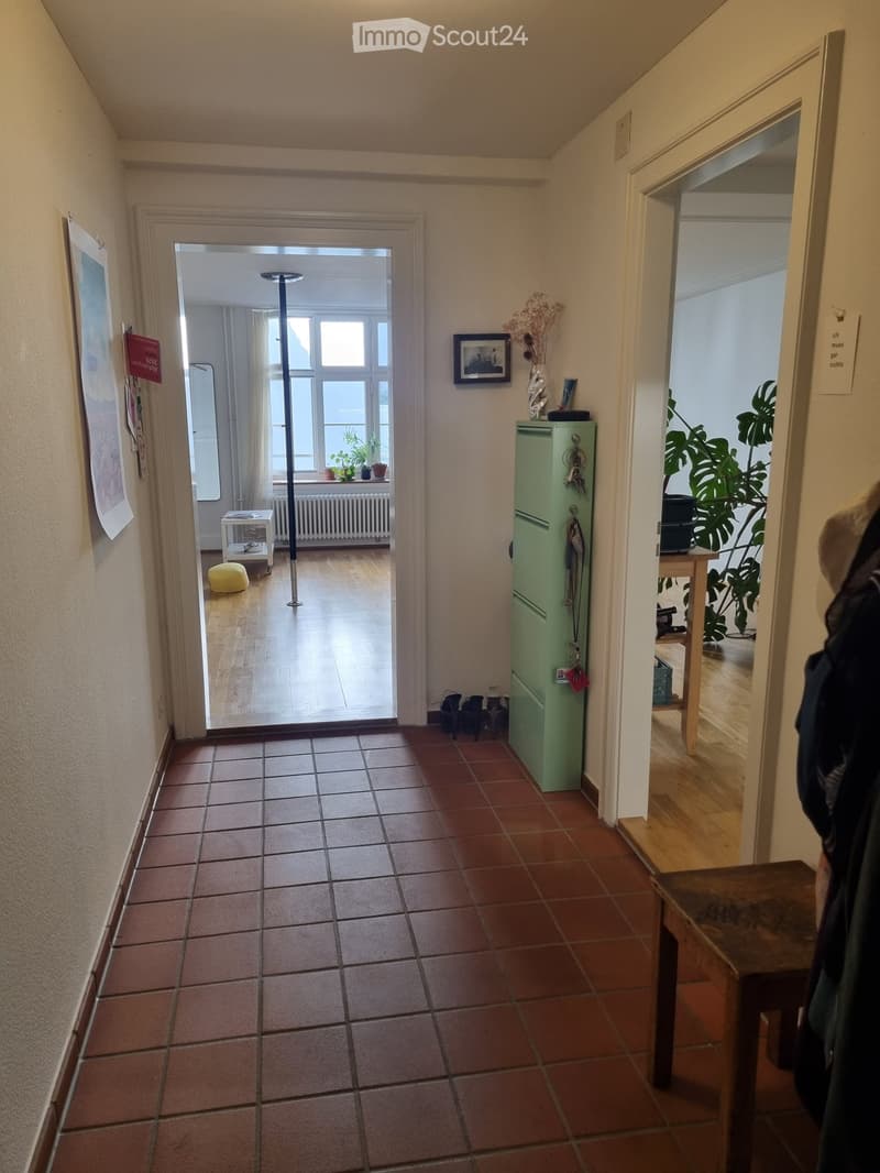 A narrow hallway with tiled floor, leading to two rooms. The left room has wooden flooring, a small table, and a plant. The right room features a green cabinet, a large plant, and a coat rack with jackets.