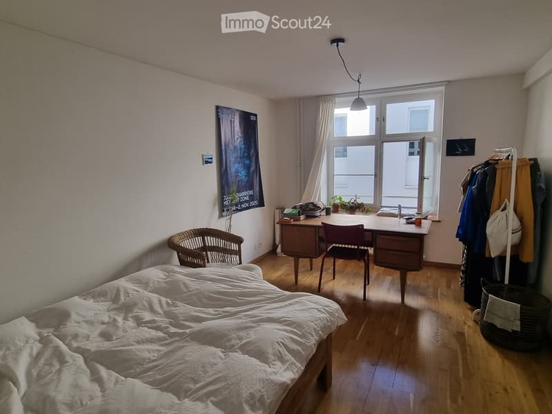 Cozy bedroom with a double bed, wooden floor, a desk with a chair, a wicker chair, a coat rack, and a poster on the wall.