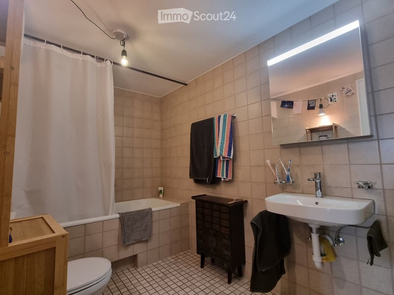 Bathroom with beige tiled walls and floor, featuring a bathtub, toilet, sink, mirror, and towel rack with towels. A shower curtain is visible in the background, and a small wooden cabinet is present.