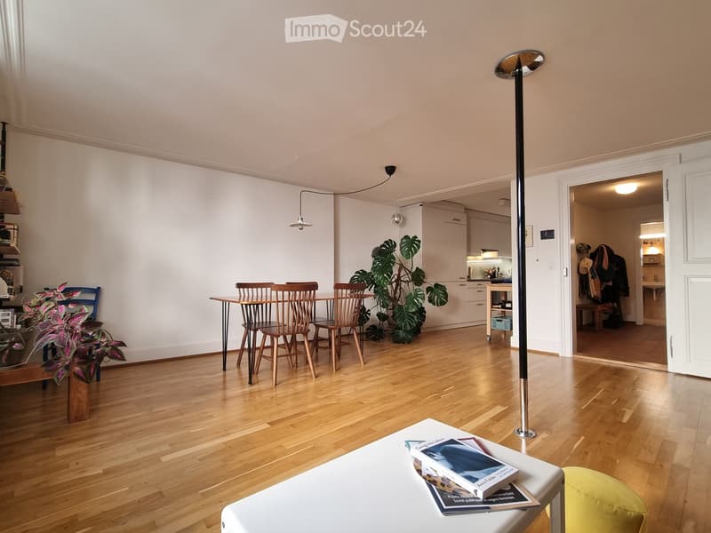 Bright living room with wooden flooring, white walls, and modern decor. Features a coffee table with books, a floor lamp, a dining area with chairs and a table, and large potted plants.
