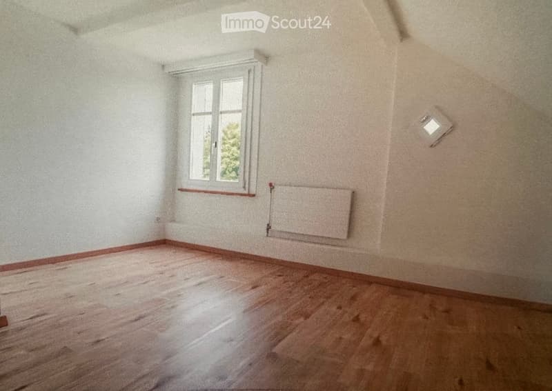 Empty room with wooden flooring, white walls, a window, and a radiator. No furniture or equipment present.