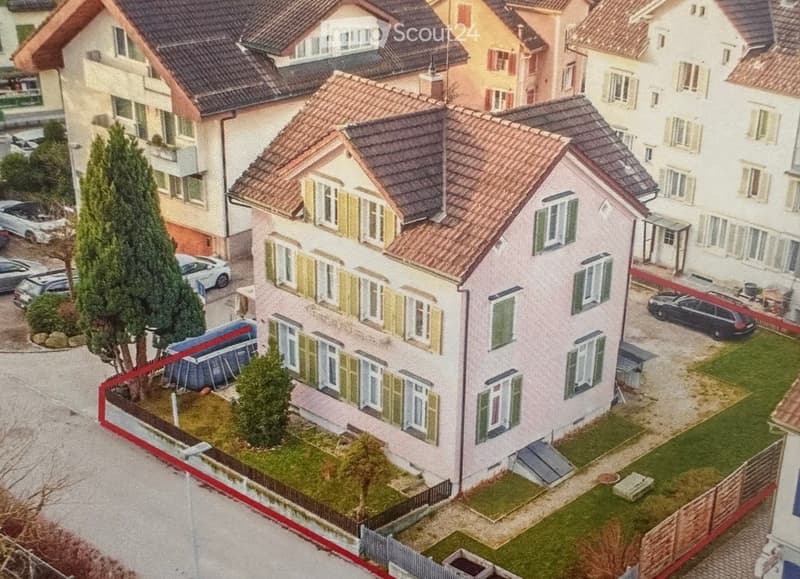 A two-story house with a red-tiled roof, multiple windows with shutters, and a small garden area with trees and a fence.
