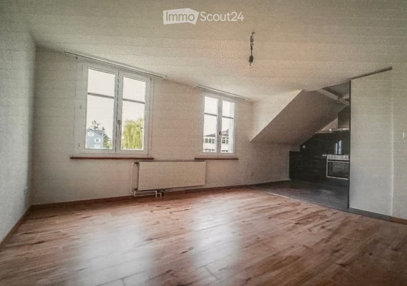 Empty room with wooden flooring, large windows, radiator, and a small kitchen area visible in the background.