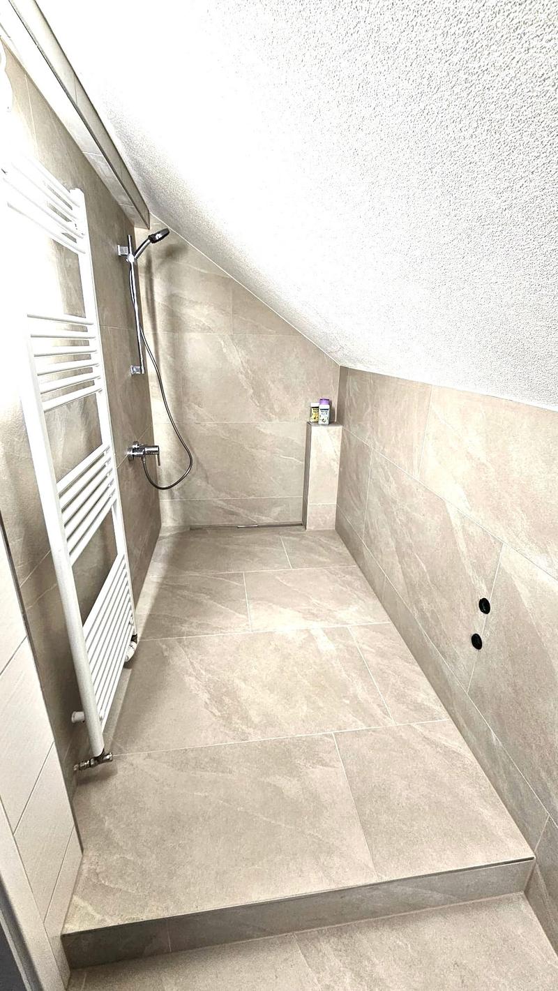 Modern shower area with beige tiles, a white radiator, and a glass shower enclosure. A small shelf holds a bottle of shower gel.