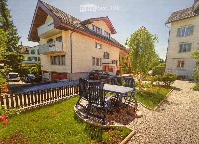 Garden area with a table and chairs, surrounded by a wooden fence, with a multi-story house and parked cars in the background.