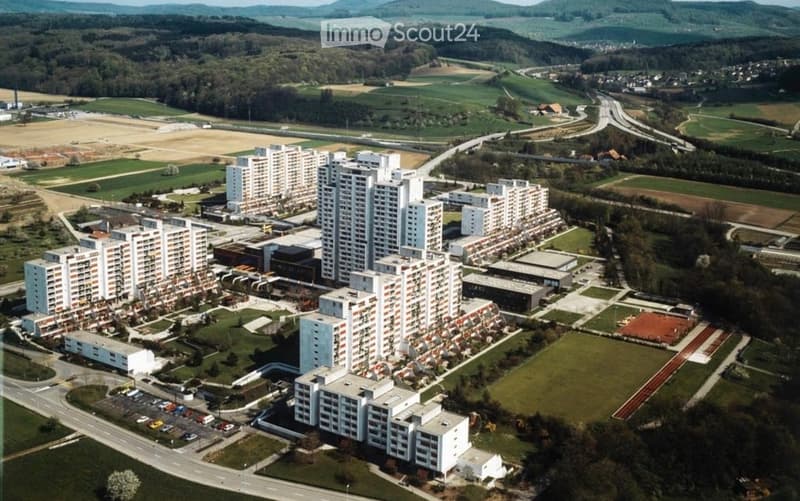 Aerial view of a large residential complex with multiple high-rise apartment buildings, surrounded by green spaces, roads, and distant hills.