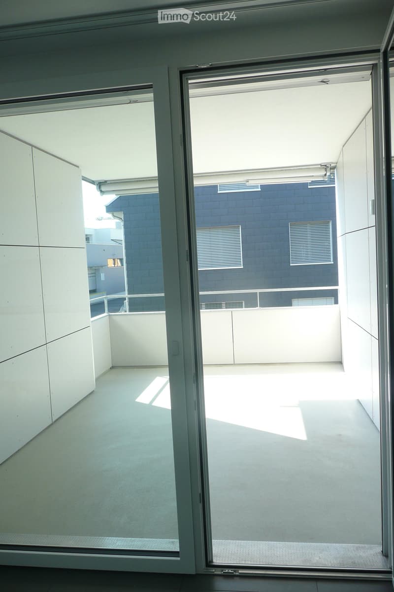 Modern balcony with large glass doors, white walls, and a view of a blue brick building with windows and vents.