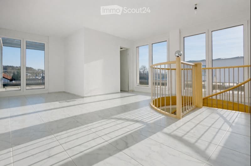 Bright, empty room with white walls, large windows, and a modern wooden staircase leading upwards.