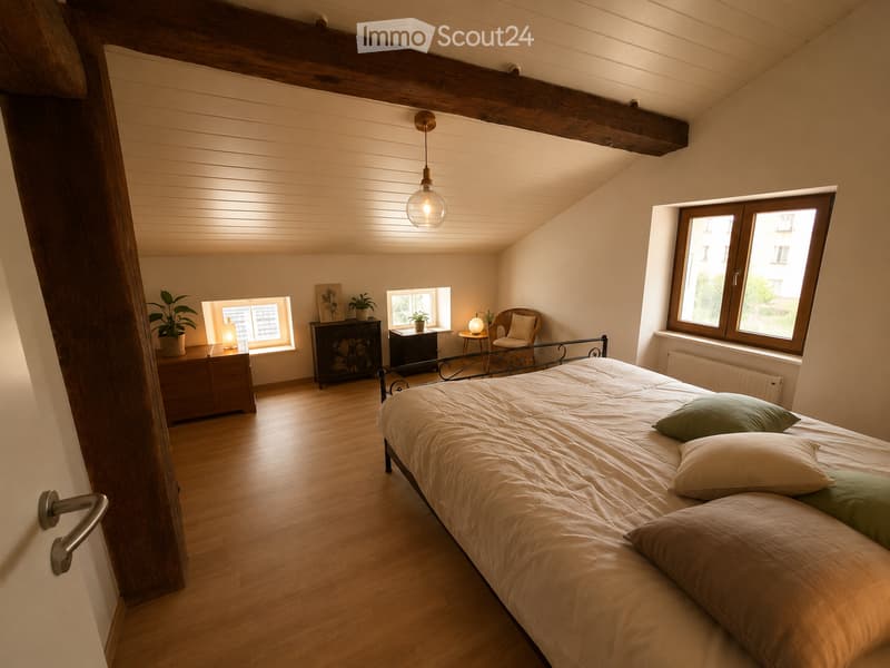 Cozy attic bedroom with wooden floors, slanted wooden ceiling beams, large windows, a neatly made bed with pillows, a nightstand, a small table with a lamp, and potted plants.