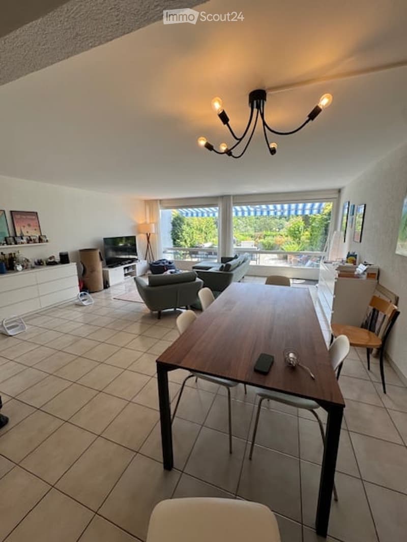 Bright, open-plan living and dining area with large windows offering a view, a wooden dining table with chairs, a grey sofa, an armchair, a TV, and a floor lamp.