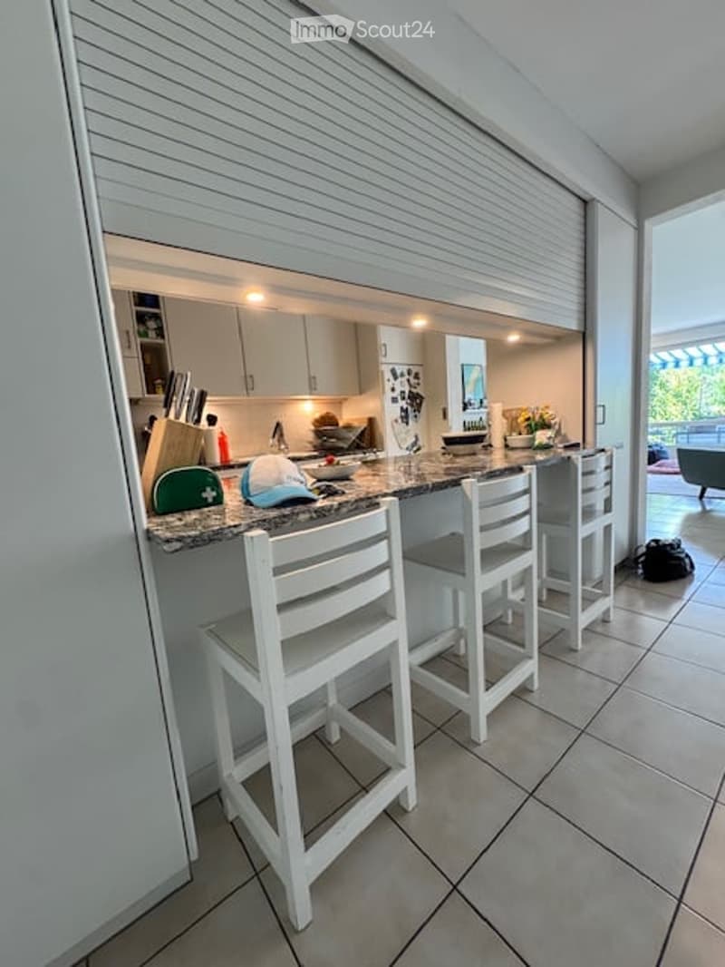 Modern kitchen with a bar counter, white stools, and a view to a balcony. The counter features a knife block, a bowl, a vase with flowers, and a mirror. The kitchen cabinets are white and sleek.