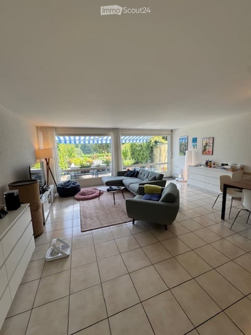 Bright living room with large windows offering a view, featuring a grey sectional sofa, coffee table, armchairs, floor lamp, TV stand, dining area with chairs and table, and white cabinets.