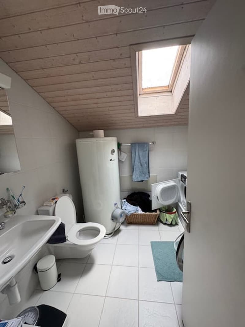 Bathroom with a skylight, white tiled floor, sink, toilet, water heater, and washing machine. A towel hangs on a rack, and a small rug is placed on the floor.