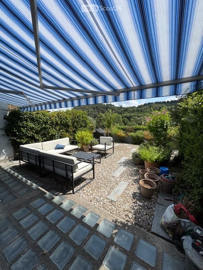 Outdoor patio area with a striped canopy, white sectional sofa, small coffee table, and potted plants. Gravel flooring and a view of greenery in the background.