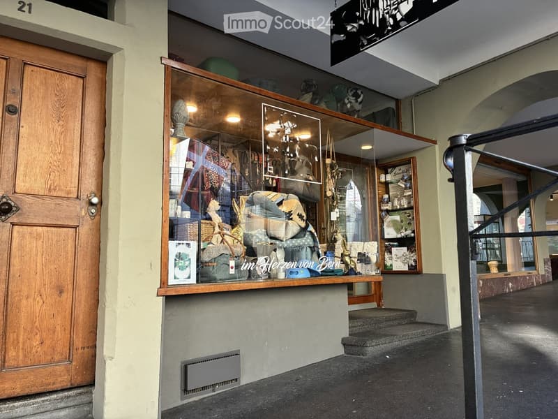 Shopfront with glass display window showcasing various items including bags, textiles, and decorative objects. Wooden door and staircase leading up to the entrance.