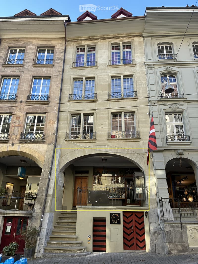 A multi-story building with ornate architecture, featuring arched entrances, balconies with metal railings, and large windows. The facade includes decorative stonework and a flag displayed on the right side.