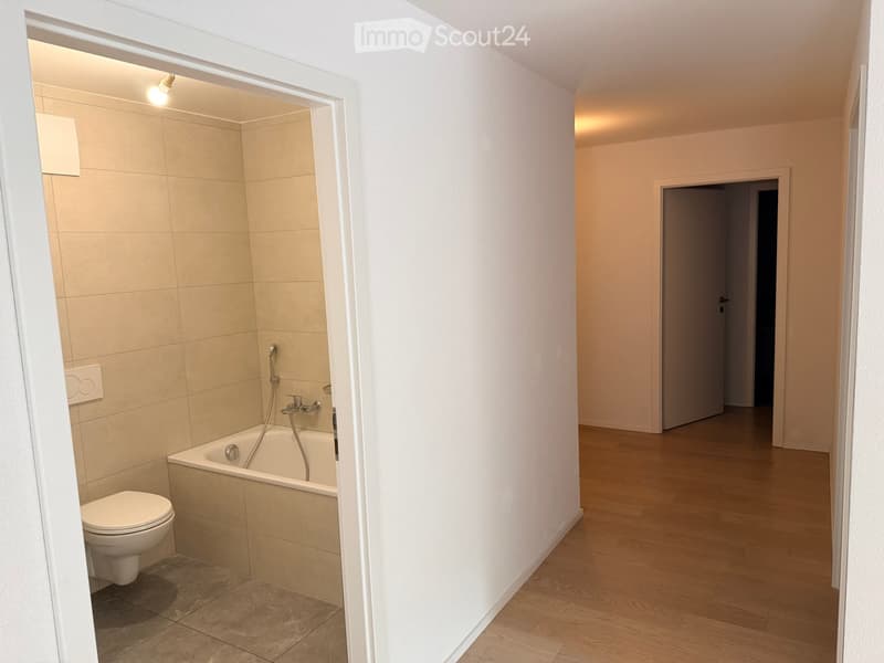 Modern bathroom with a toilet, bathtub, and a sleek faucet. The room features beige tiles and warm lighting.