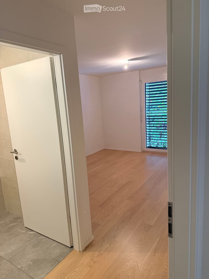 Empty room with light wooden flooring, white walls, and a large window with blinds. The room is connected to a hallway with a white door.