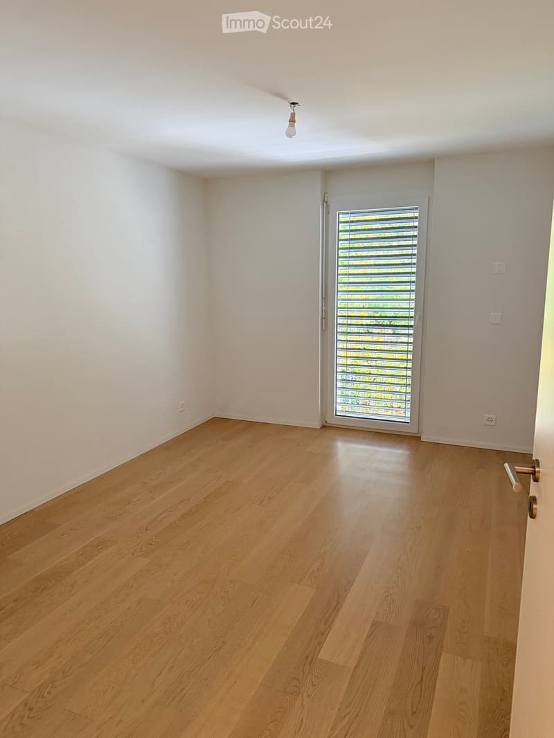 Empty room with light wooden flooring, white walls, and a sliding glass door with blinds.