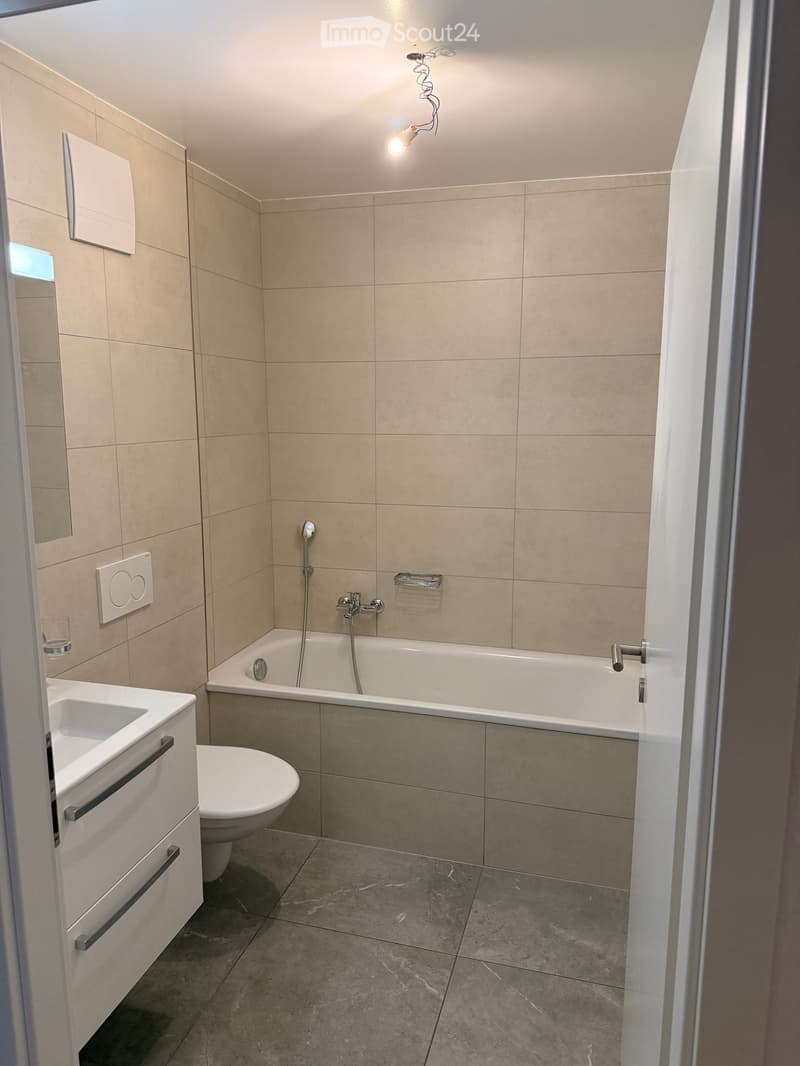 Modern bathroom with beige tiled walls, a white sink, toilet, bathtub, and a mirror. Features a sleek faucet and a light fixture above the bathtub.