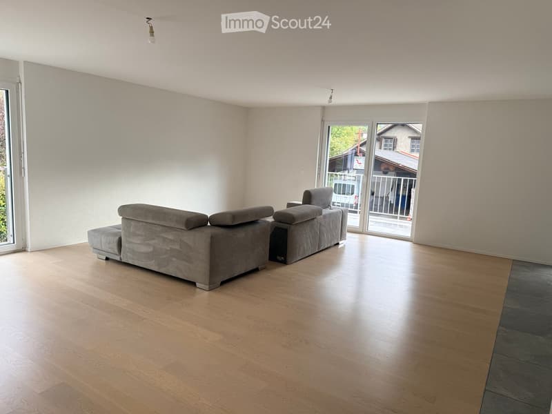 Modern living room with light wooden flooring, grey sectional sofa, large windows, and sliding glass doors leading to a balcony.