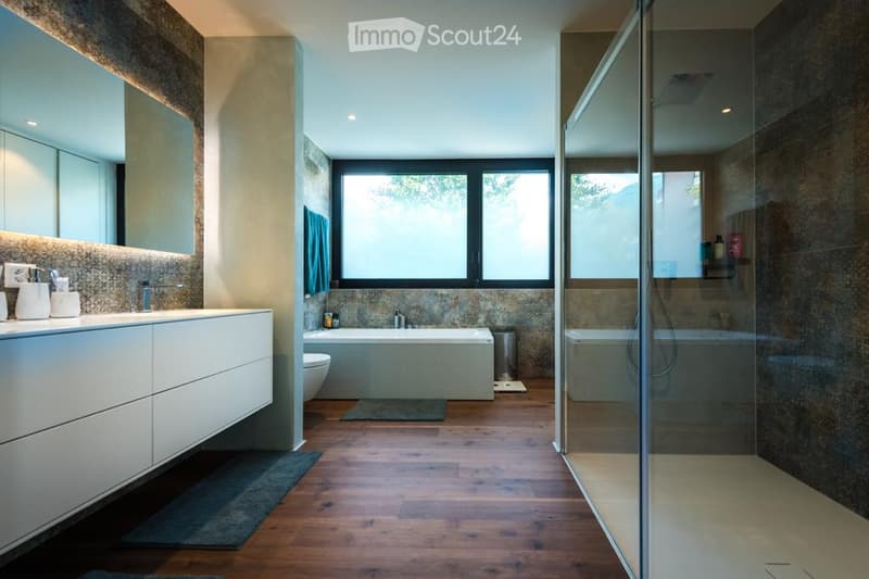 Modern bathroom with a large window, white sink, mirror, bathtub, toilet, and a glass-enclosed shower. Wooden flooring and natural light.