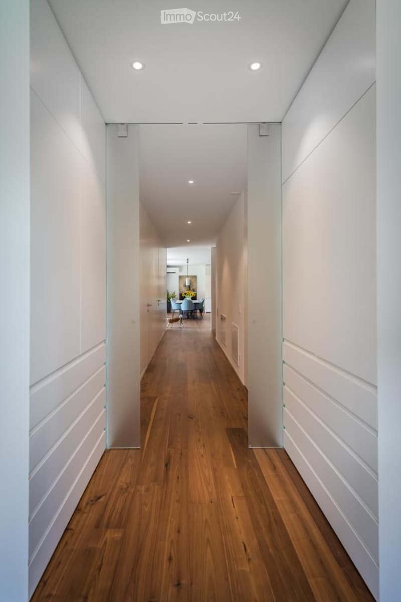 Modern hallway with white walls, wooden flooring, recessed lighting, and a sliding glass door leading to a dining area.