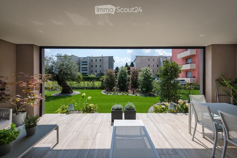 Spacious balcony with wooden flooring, modern furniture including chairs and a table, surrounded by greenery and a view of a landscaped garden and buildings.