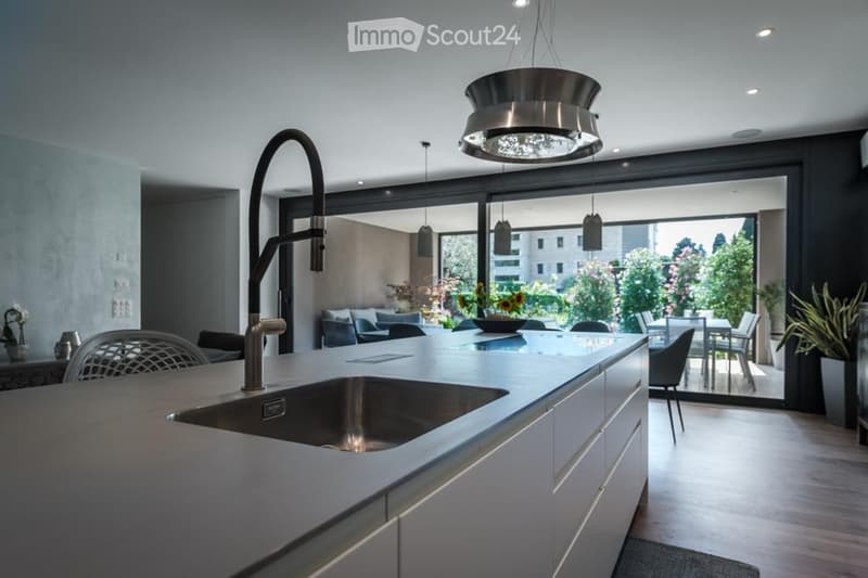 Modern kitchen with a central island, stainless steel sink, sleek faucet, pendant lights, and a dining area visible through large windows.