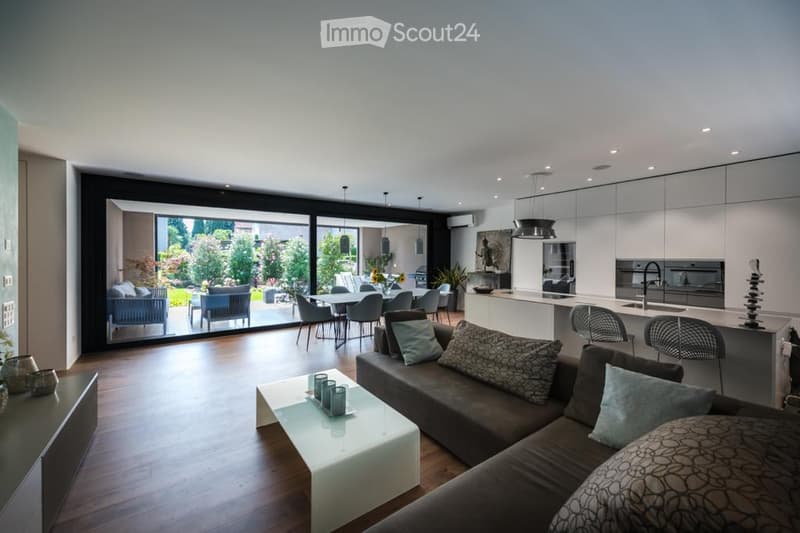 Modern open-plan living and dining area with large glass doors leading to a garden, featuring a grey sectional sofa, white coffee table, dining table with chairs, and a sleek kitchen island with pendant lights.