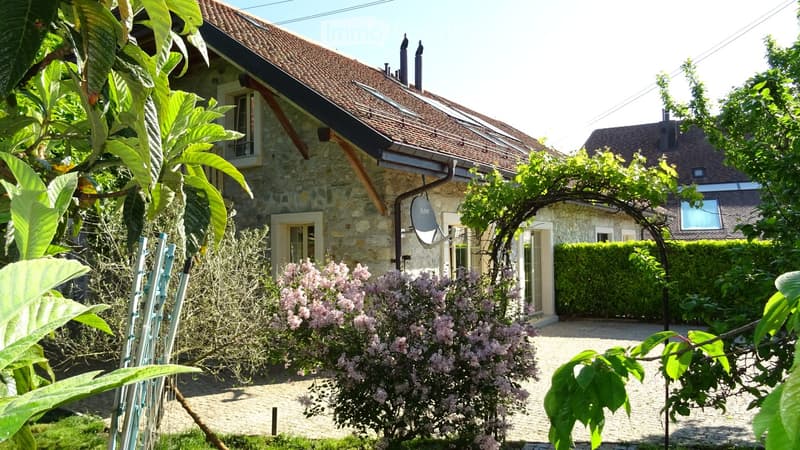 A stone house with a tiled roof, surrounded by a garden with flowering shrubs and trees. The garden features a gravel pathway and a small pergola structure.