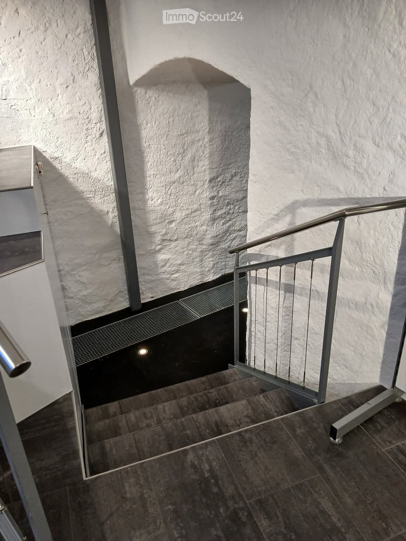 A modern staircase with a metal railing and tiled steps, leading upwards with a textured white wall and a small light fixture visible in the background.