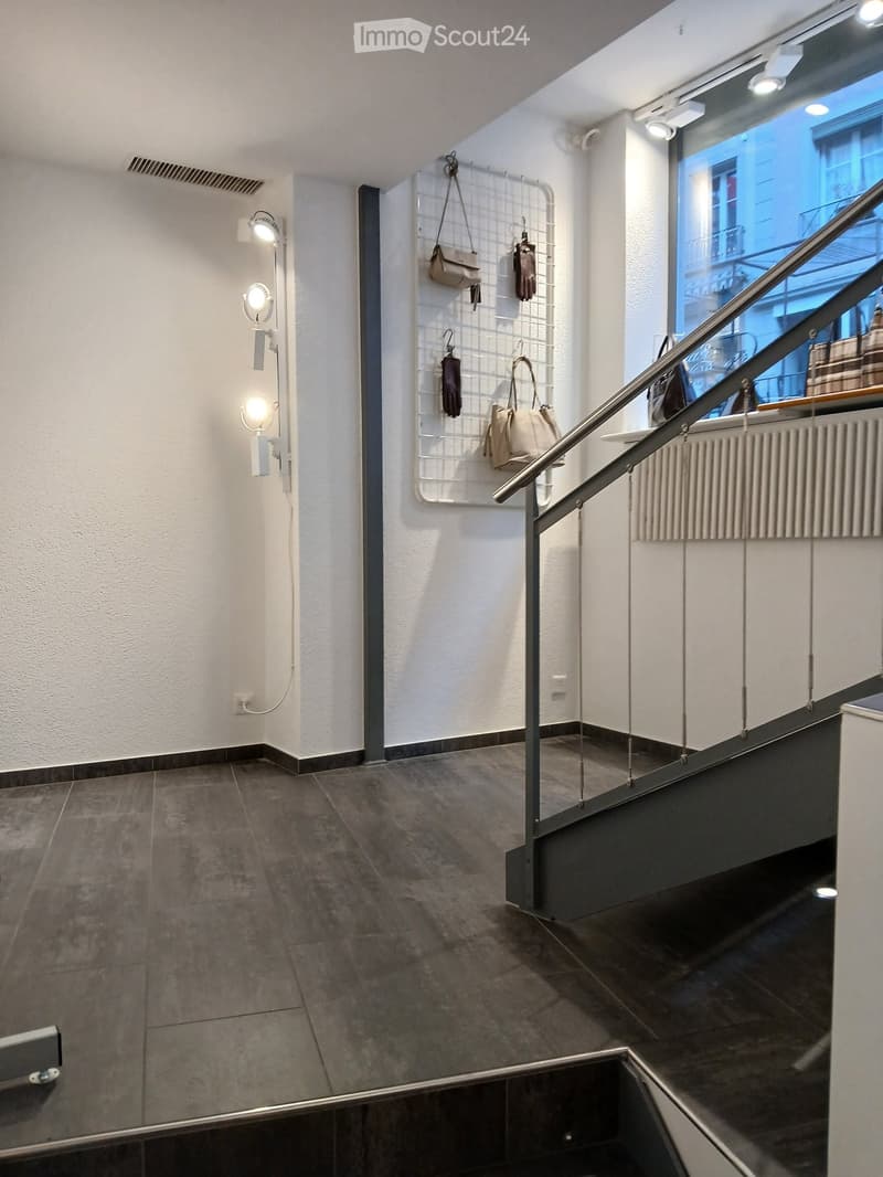 Modern hallway with tiled floor, white walls, and a staircase leading upwards. A coat rack with several bags and coats is mounted on the wall, and modern wall-mounted lighting fixtures are visible.