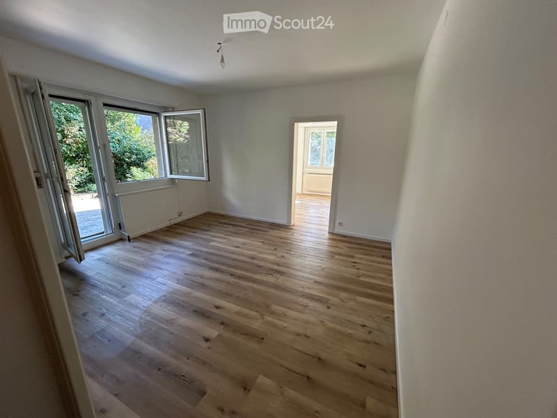 Empty room with light wooden flooring, white walls, and large windows with open sliding doors leading to an outdoor area.