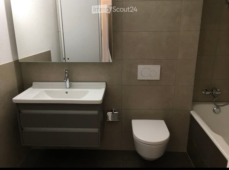 Modern bathroom with a white sink, chrome faucet, large mirror, toilet, and bathtub. Beige tiled walls and dark flooring.