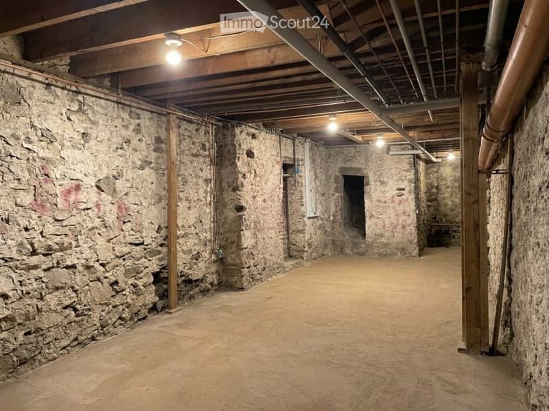Empty basement with exposed stone walls, wooden support beams, and exposed pipes. The floor is concrete, and the ceiling has exposed wooden beams and lighting fixtures.