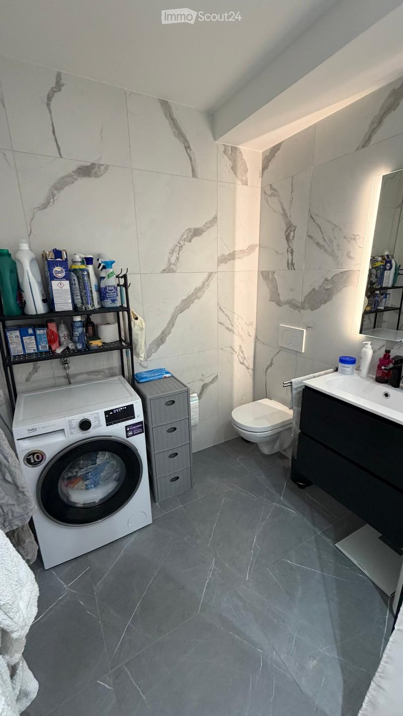 Modern bathroom with marble tiles, a washing machine, toilet, sink, and shelves holding cleaning supplies.