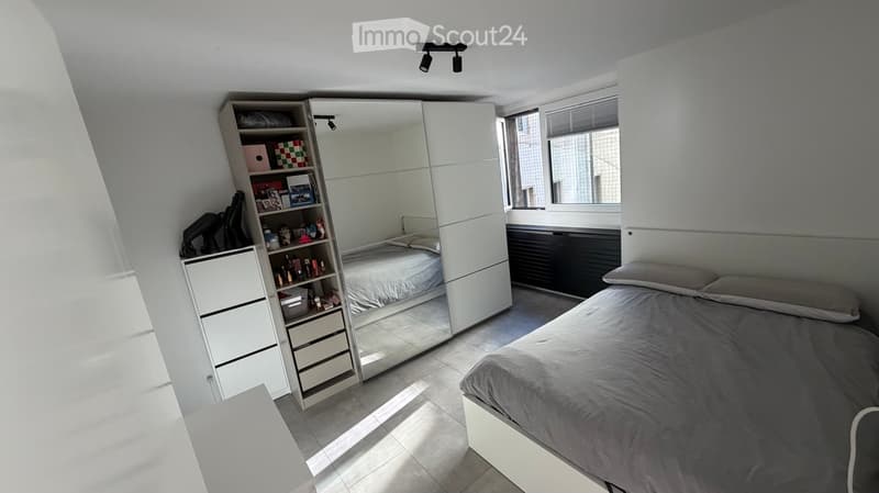 Modern bedroom with a large bed, mirrored wardrobe, built-in shelving unit, and a window with blinds.