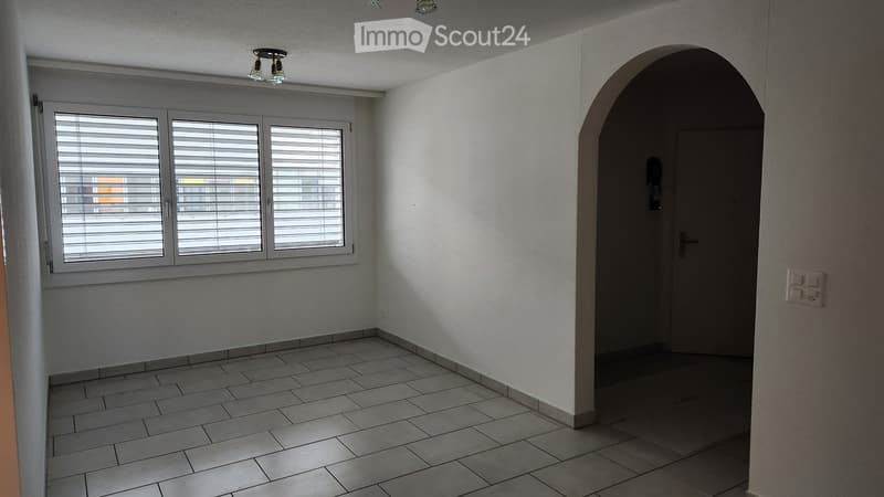 Empty room with tiled floor, white walls, large window with blinds, and an arched doorway leading to another space.