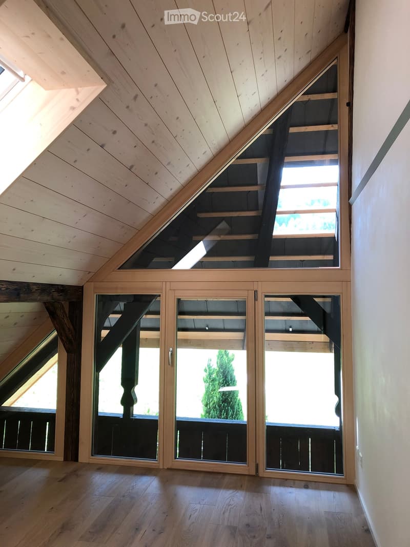 An empty attic space with wooden floors, exposed wooden beams, and large windows offering a view of the outdoors.