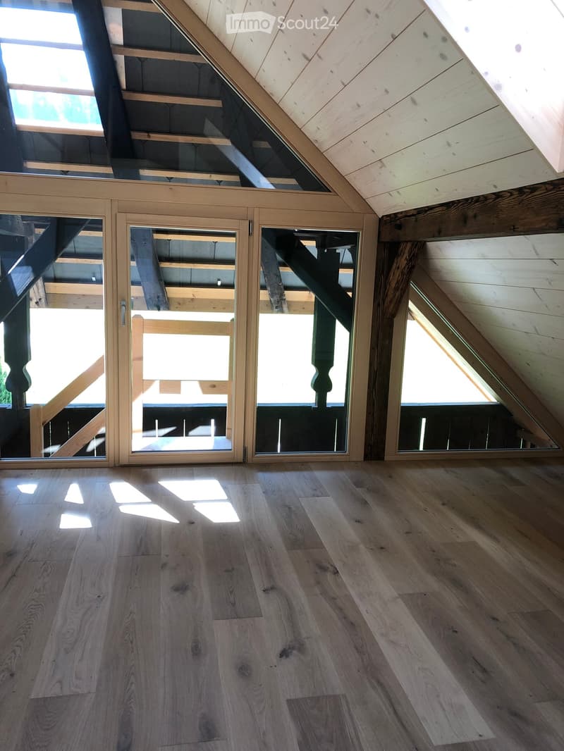 Bright, empty attic space with wooden flooring, exposed wooden beams, and large glass doors leading to a balcony or outdoor area.