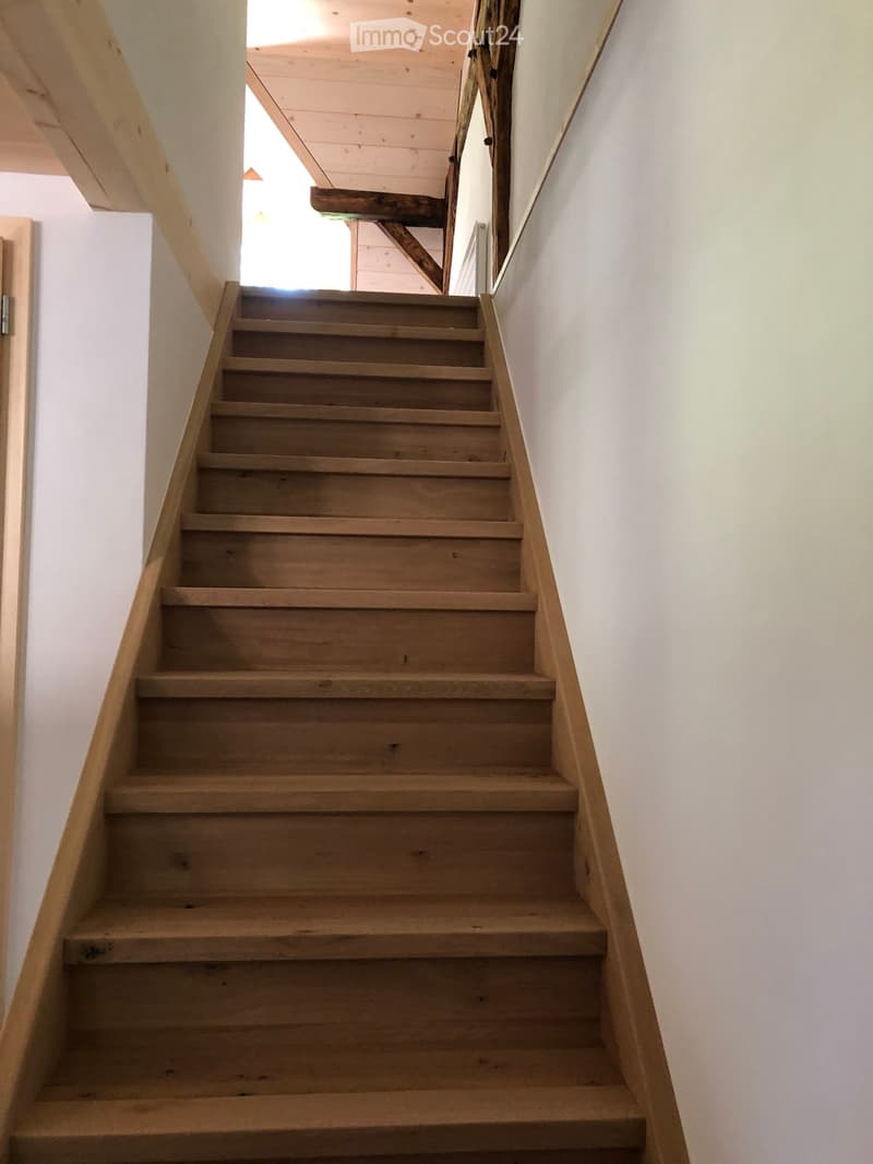 Wooden staircase with a wooden handrail, white walls, and a visible ceiling with exposed wooden beams.